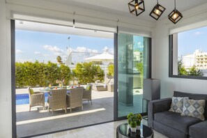 A modern living room with a grey sofa, a glass door leading to a balcony with outdoor furniture and a pool. - Pernera Beach Villa in Protaras (Paralimni)