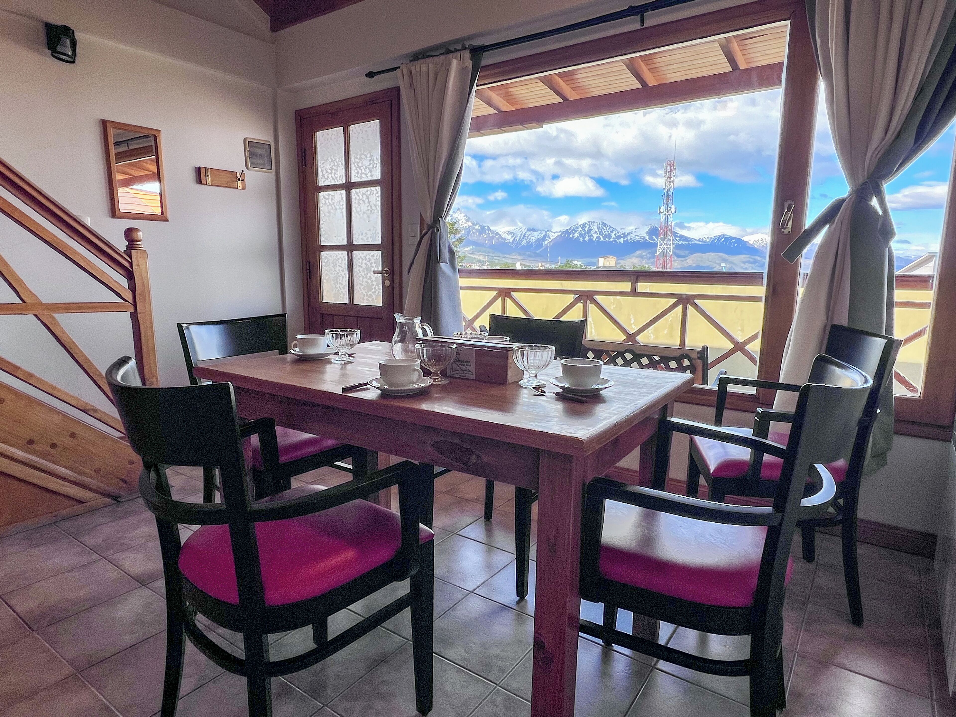 Zona pranzo con tavolo e sedie in legno, vista sulle montagne attraverso una finestra e una scala a sinistra.