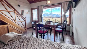 A room with a bed, a dining table with chairs, a staircase, and a view of mountains through a window.