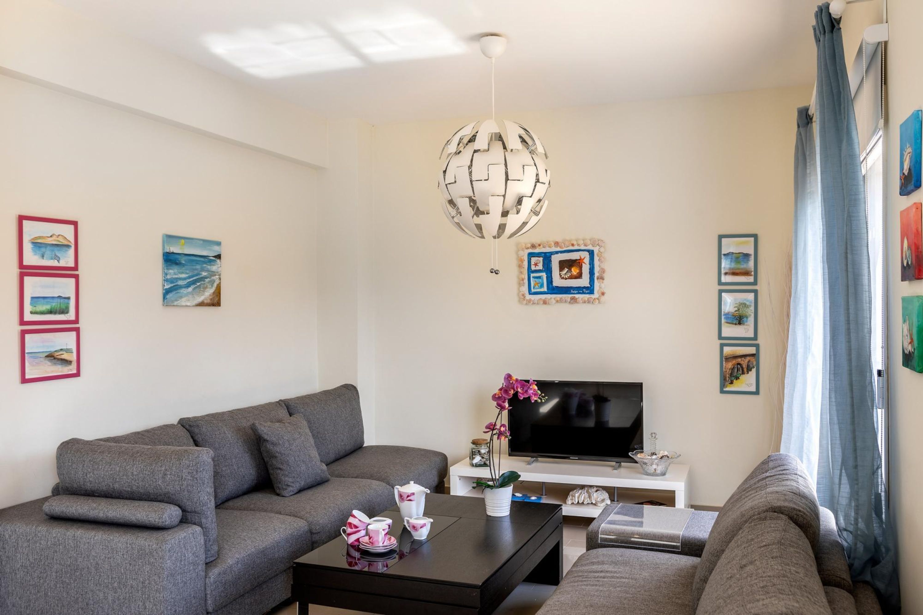 A modern living room with a grey sofa, a coffee table, and a flat-screen TV. There are framed artworks on the walls and a globe light fixture.