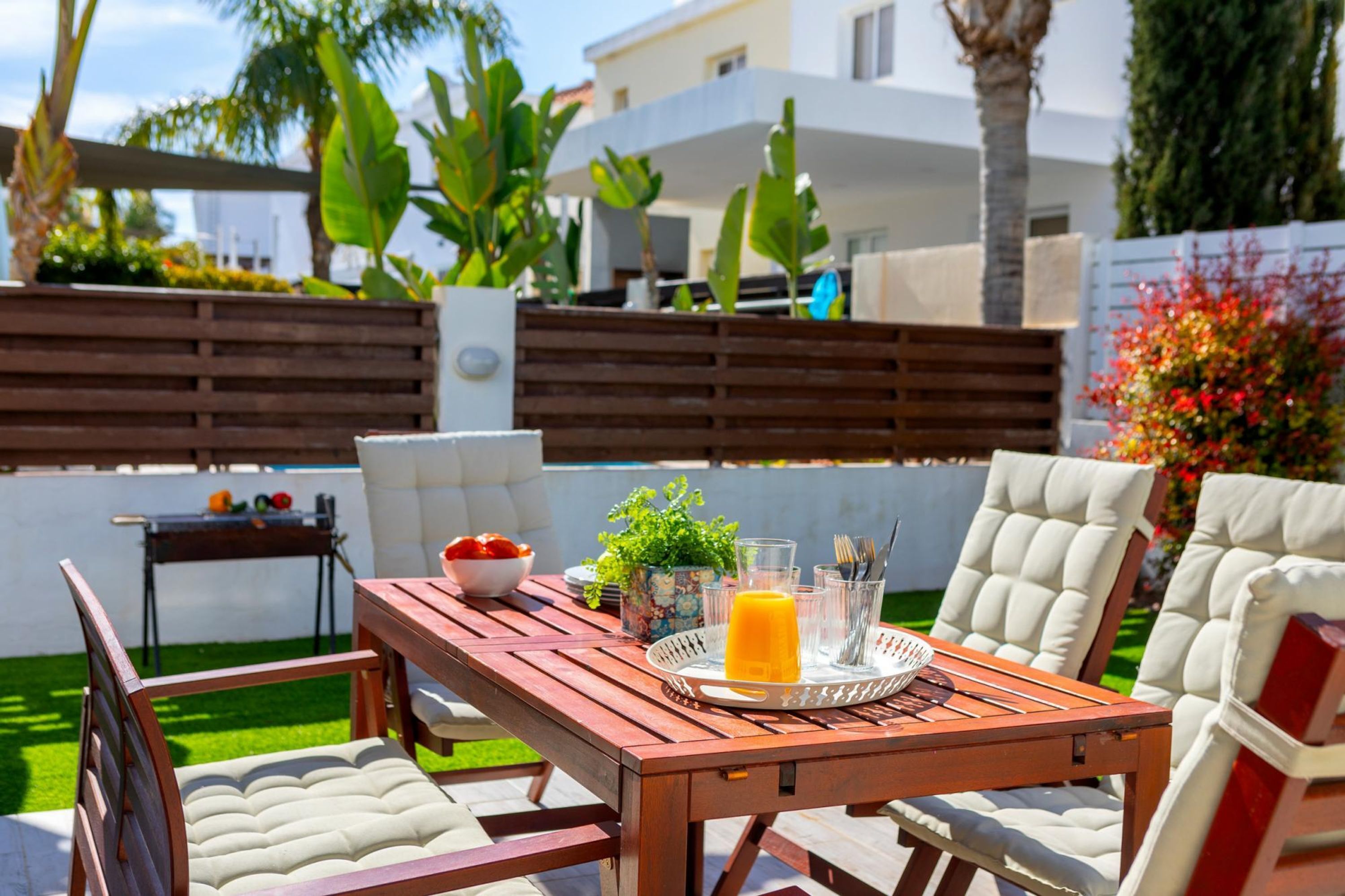 An outdoor patio with a wooden table, white cushioned chairs, and a small garden area with plants and a fence.