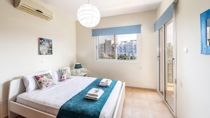 A bedroom with a bed, a window with a view of buildings, a ceiling fan, and an air conditioning unit. - Galleria Sunset Villa (Paralimni)