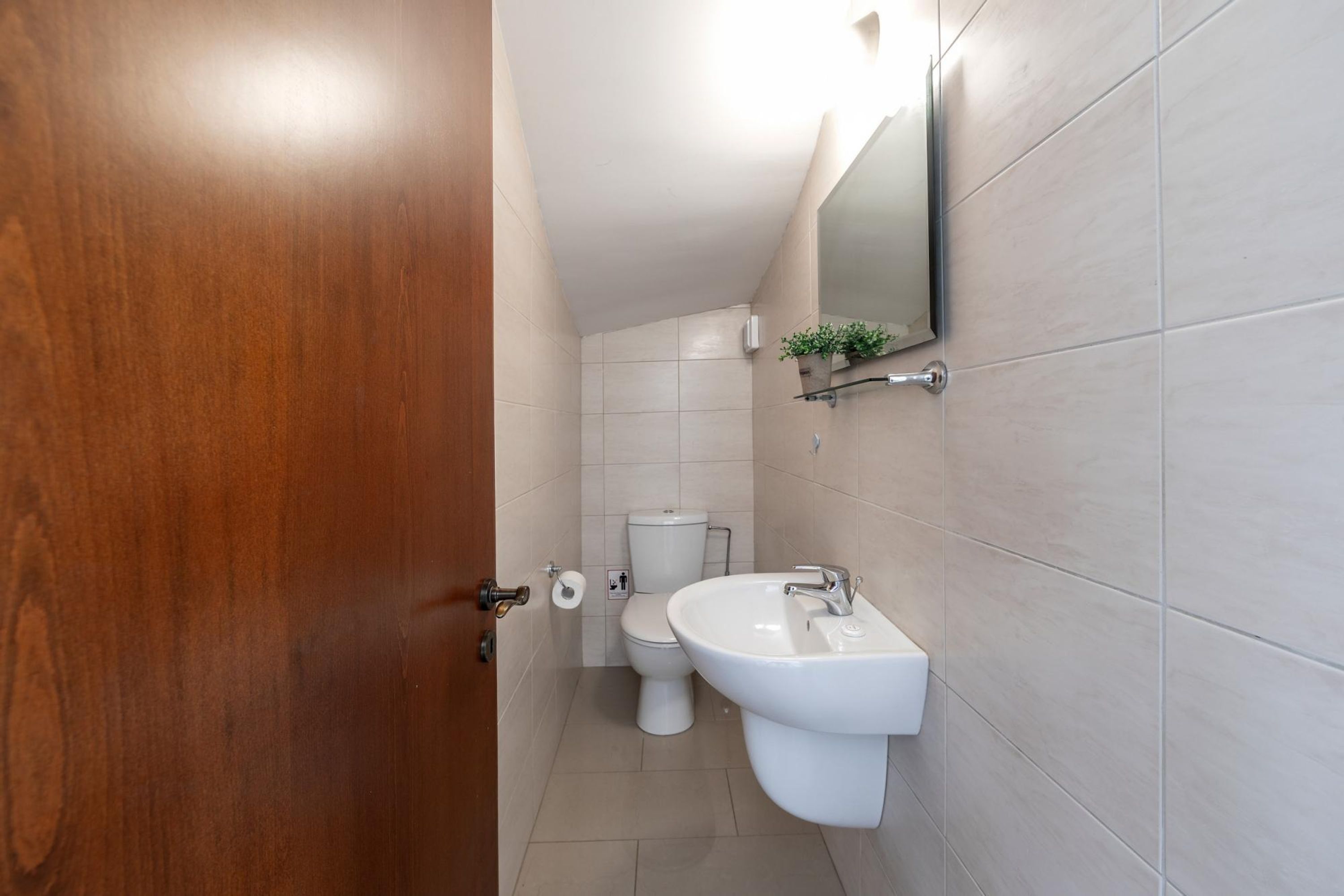 Une salle de bain avec une porte en bois, des toilettes blanches et un lavabo surmonté d’un miroir.