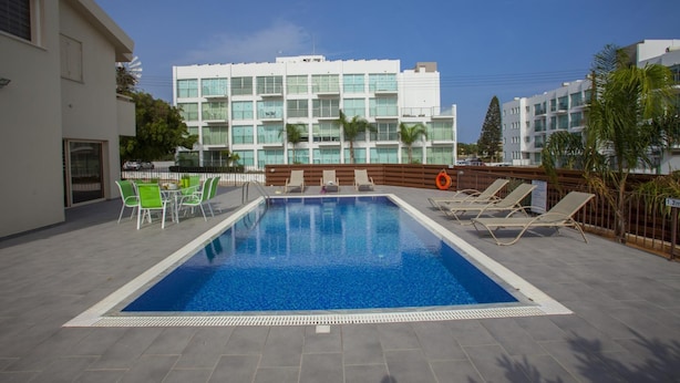 A swimming pool with lounge chairs and a table, surrounded by a modern building with multiple floors and large windows. - Theodora 16 in Protaras (Paralimni)