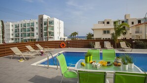 A rooftop pool area with lounge chairs, a table with breakfast items, and a view of the sea and buildings. - Theodora 16 in Protaras (Paralimni)