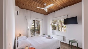 Une chambre avec un plafond en bois, un ventilateur de plafond, un lit avec du linge de lit blanc, un téléviseur fixé au mur et une fenêtre donnant sur les arbres.
