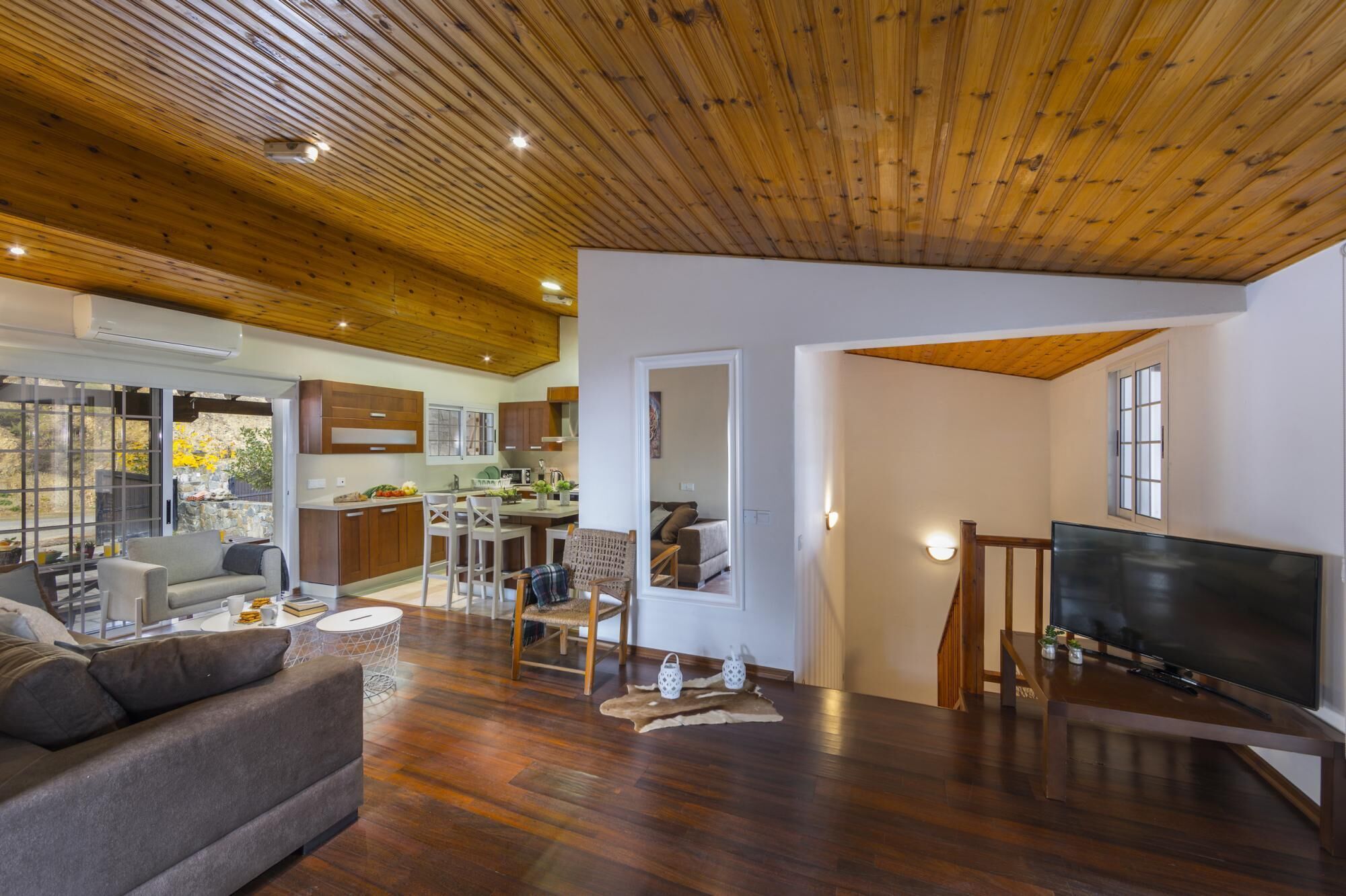 A spacious living area with a wooden ceiling, a sofa, a TV, and a kitchen in the background.