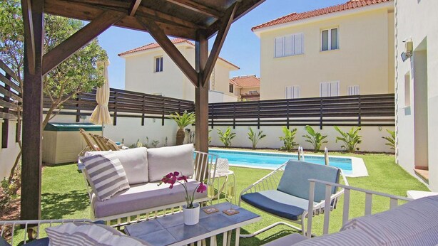 Un espace extérieur aménagé avec une zone de repos couverte, une piscine, une clôture blanche et un bâtiment en arrière-plan.