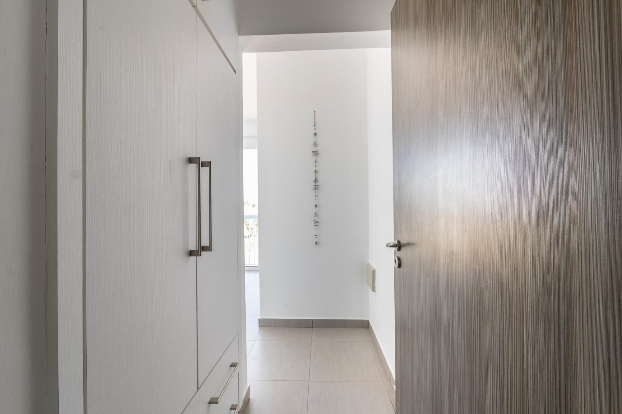 Un couloir avec une porte en bois, un mur blanc orné d’un motif à rayures verticales et un sol carrelé.