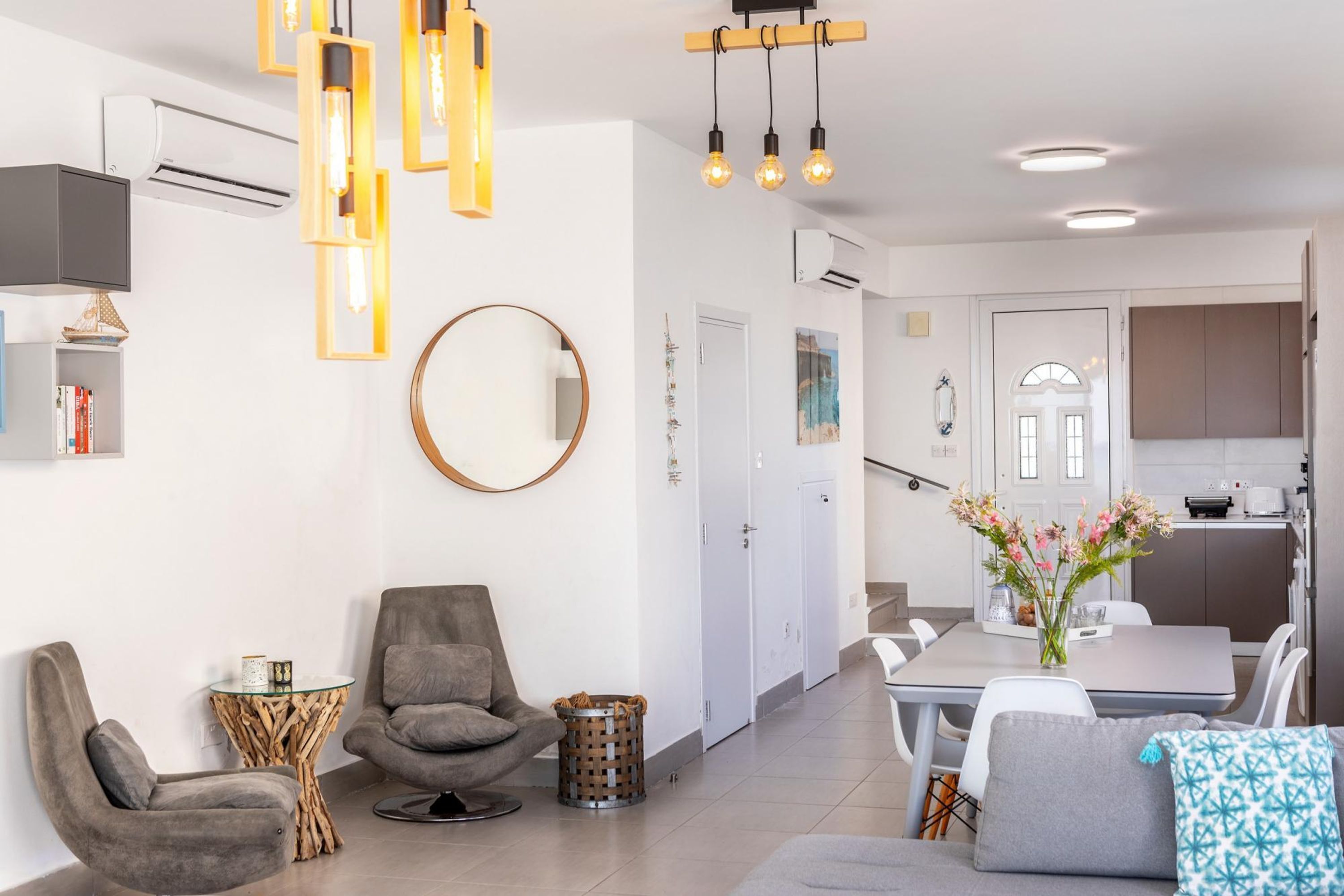 Un salon moderne avec un canapé, un fauteuil et une table à manger avec des chaises. On y trouve un miroir rond accroché au mur, une petite table d’appoint et une poubelle. La pièce est décorée dans des tons neutres, avec des murs blancs et un canapé gris.