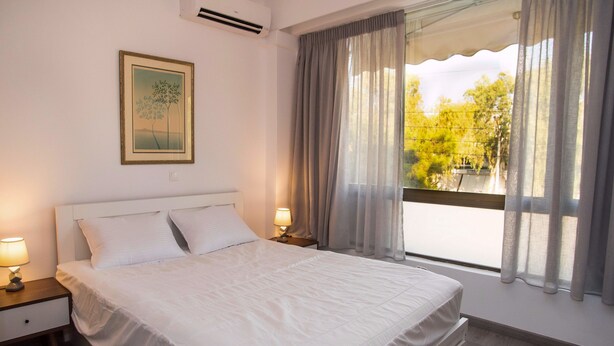 A neatly made bed in a hotel room with a window, a bedside lamp, and a framed picture on the wall. - Cozy Glyfada Getaway 1 bdr (Glyfada)