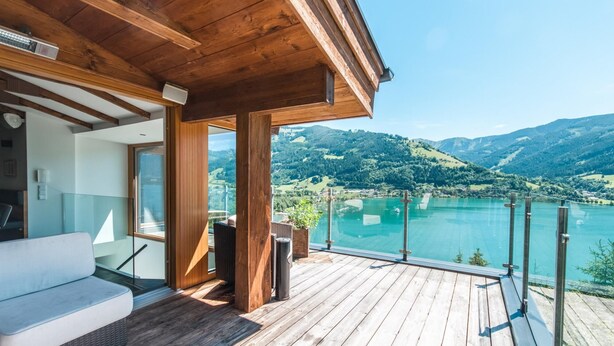Une terrasse en bois avec un canapé blanc, une balustrade en verre et une vue sur un lac et des montagnes.