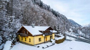 Ein gelbes Chalet mit Balkon, umgeben von schneebedeckten Bäumen und einem Berg im Hintergrund.