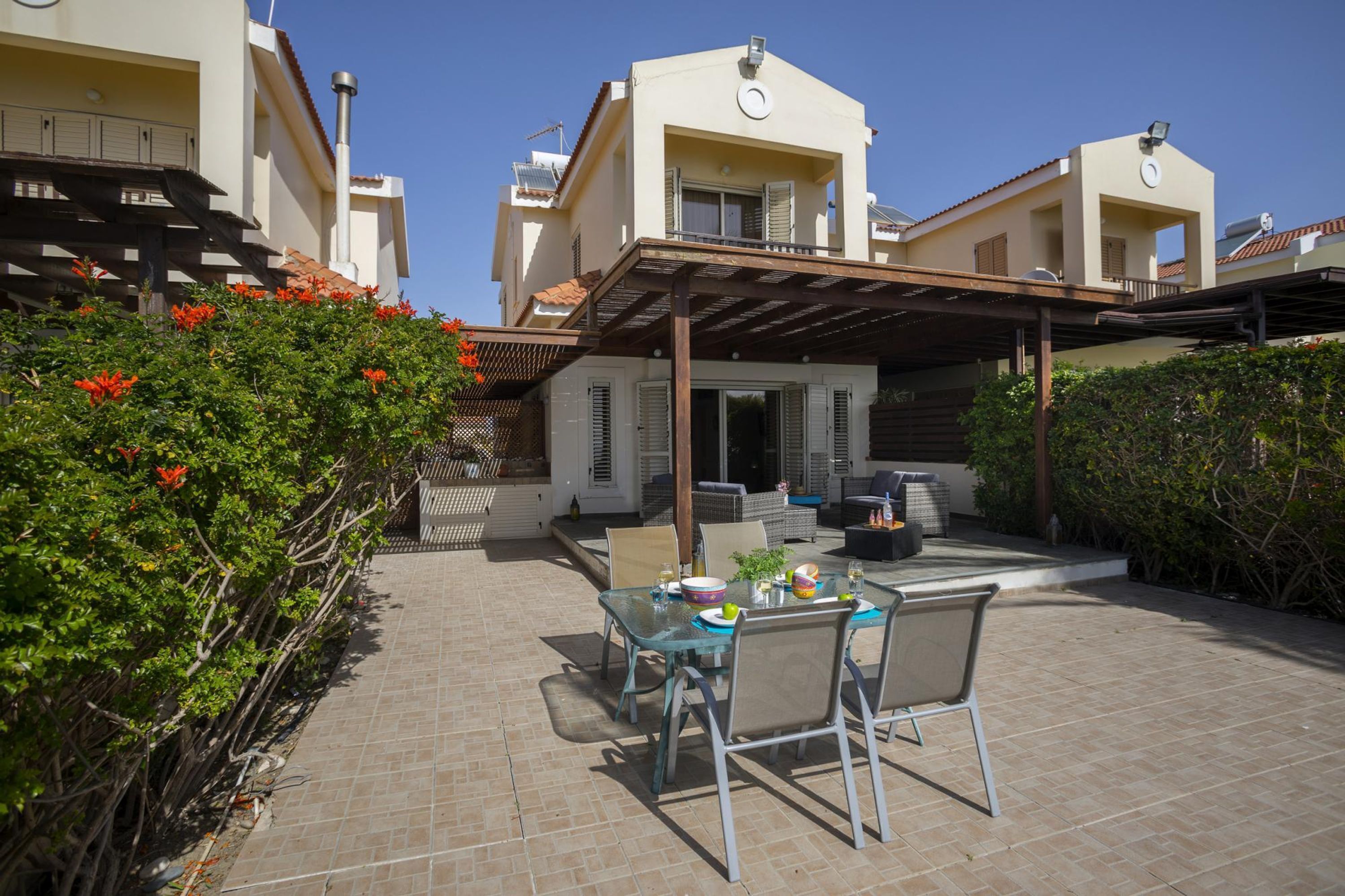 A patio with a table and chairs, surrounded by greenery and a building with a tiled roof.