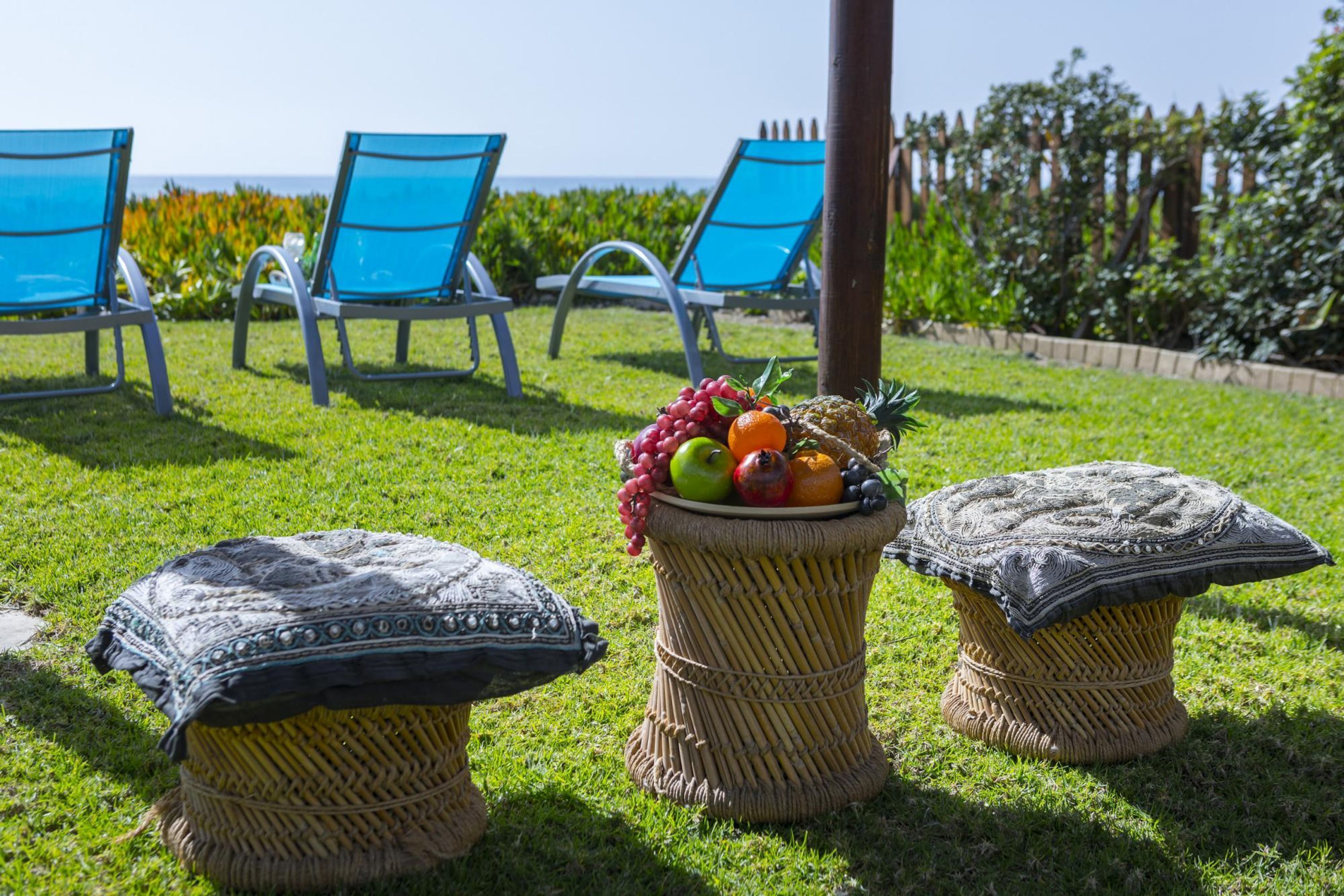 A grassy outdoor area with three blue chairs, two woven stools with cushions, and a fruit basket on one of the stools.