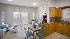 Una sala moderna con un sofá blanco, una mesa de centro y un comedor con mesa y sillas. La cocina cuenta con gabinetes de madera y una ventana con cortinas.