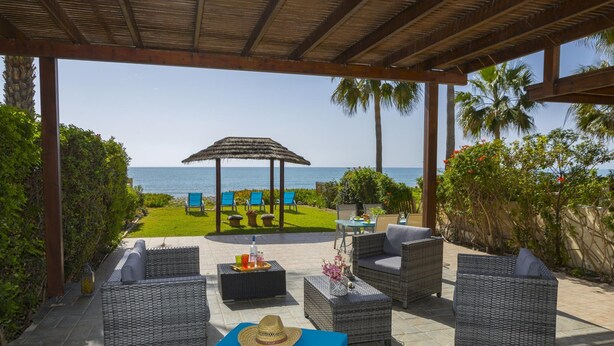 Un patio al aire libre con mobiliario de mimbre, un quiosco de paja y vista al mar.