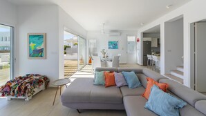 A modern living room with a grey sofa, colorful cushions, and a small round table. There is a balcony with a view of the outside. - Protaras Villa Oceania (Paralimni)