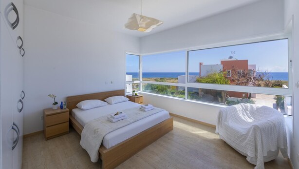A modern bedroom with a large window, a bed, a bedside table, and a nightstand. - Protaras Villa Oceania (Paralimni)