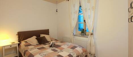 A bedroom with a wooden floor, a bed with a quilt, a nightstand, a window with curtains, and a radiator.