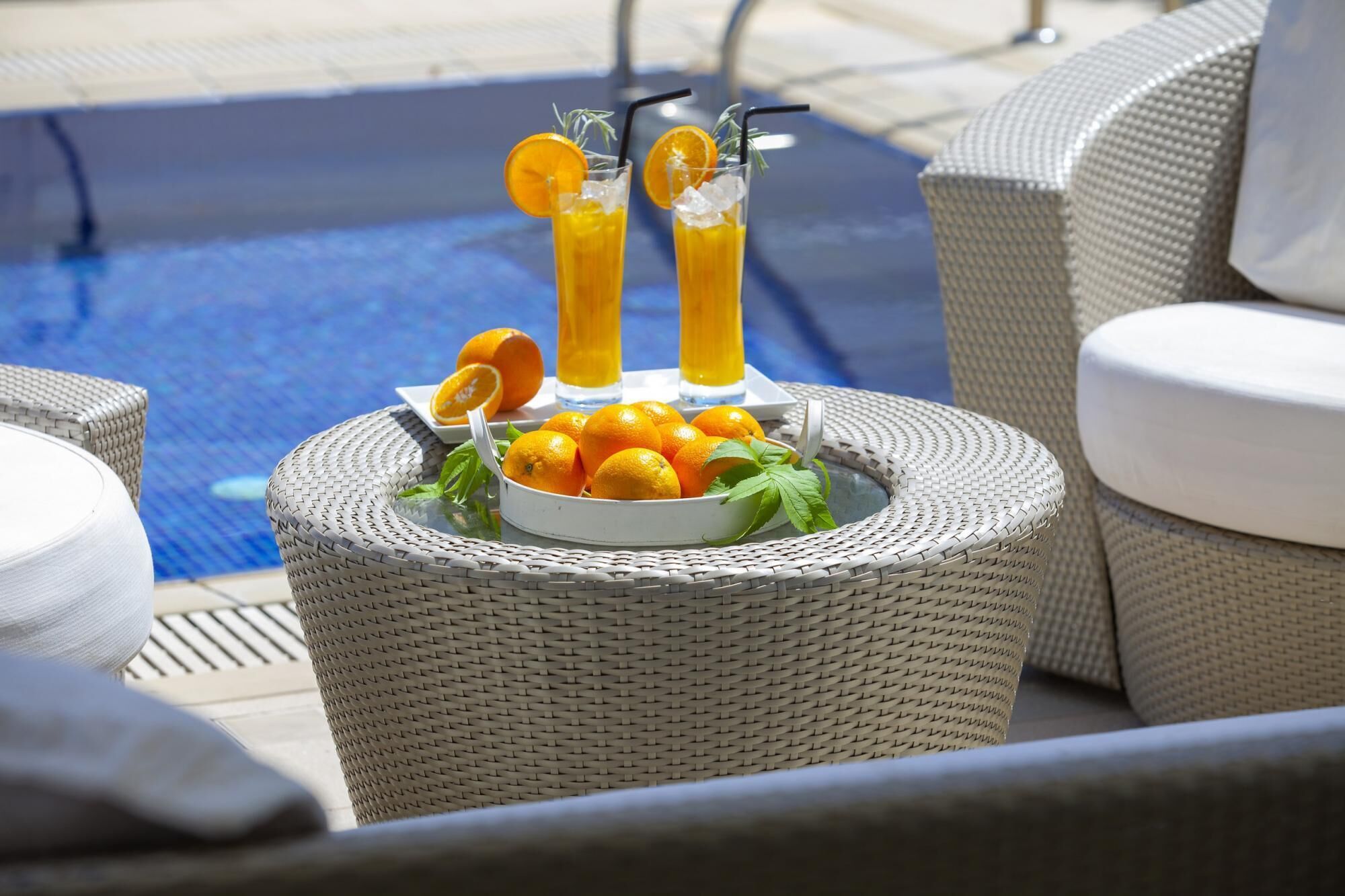A wicker table with a bowl of oranges and two glasses of orange juice, placed near a poolside chair.