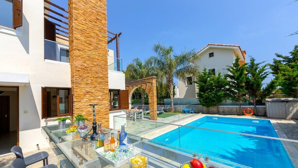 A modern outdoor pool area with a glass balustrade, a dining table, and a variety of fruits and beverages on the poolside. - Ayia Napa Ionion Seaside Villa (Sotira)