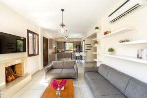 A modern living room with a fireplace, grey sofas, a wooden coffee table with flowers, and a kitchen in the background. - Ayia Napa Ionion Seaside Villa (Sotira)