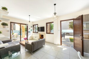 A modern living room with a fireplace, a sofa, a coffee table, and a TV. There are potted plants on shelves and a view of the outside. - Ayia Napa Ionion Seaside Villa (Sotira)