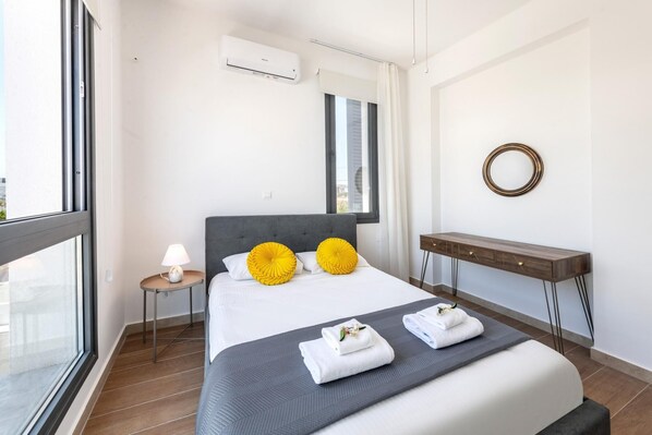 A modern hotel room with a large bed, a bedside table with a lamp, a small round table, and a mirror on the wall.