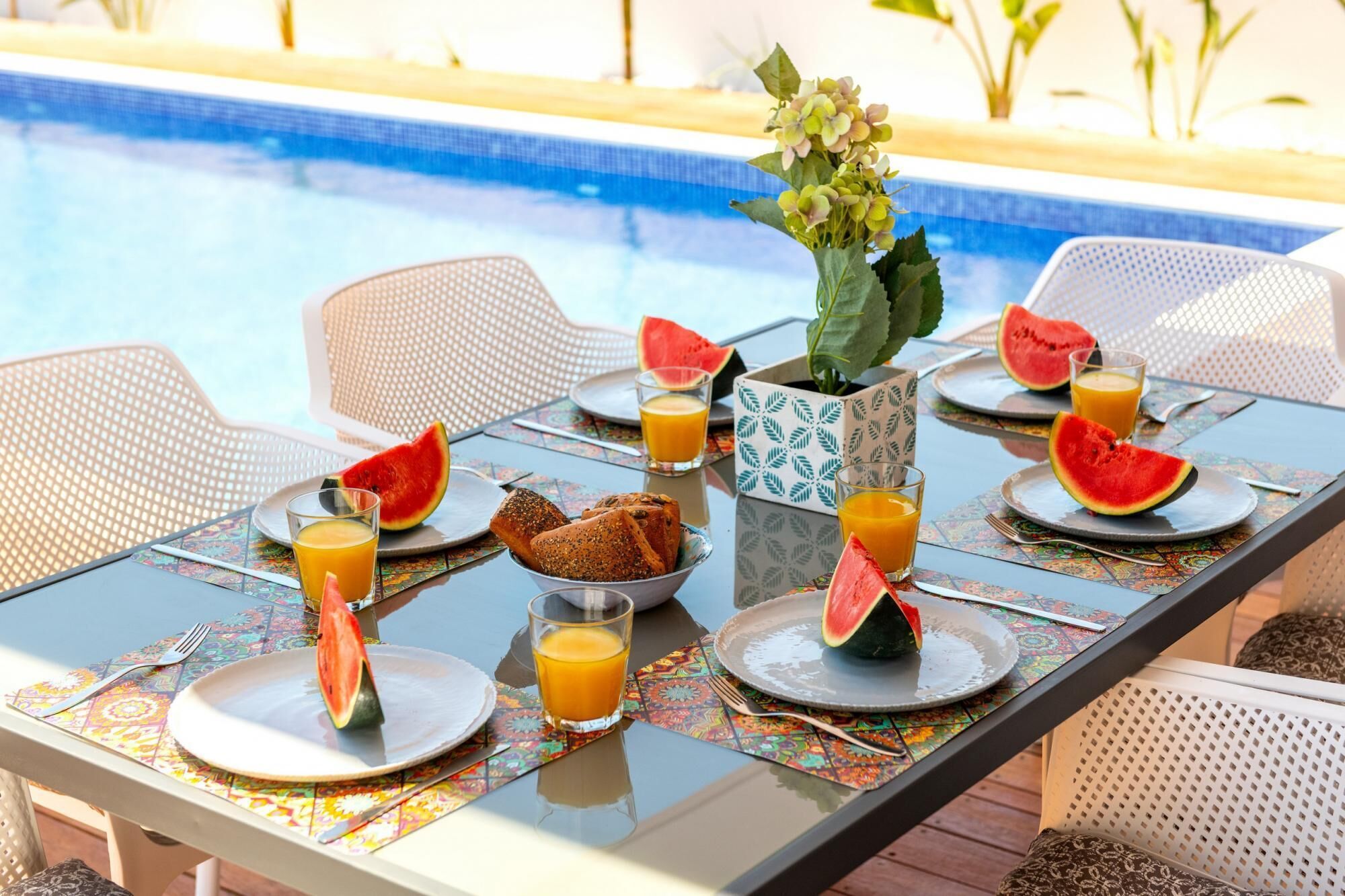 Una mesa de comedor dispuesta con platos, cubiertos, un bol de pan, vasos de zumo de naranja y rebanadas de sandía junto a una piscina.