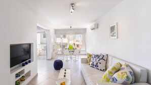 A modern living room with a flat-screen TV, a coffee table with decorative items, a sofa with patterned cushions, and a dining area in the background. - Protaras Pallini Seaside Suite (Paralimni)