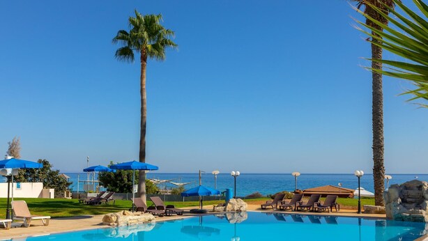 A pool area with lounge chairs and umbrellas, a palm tree, and a view of the sea. - Protaras Pallini Seaside Suite (Paralimni)