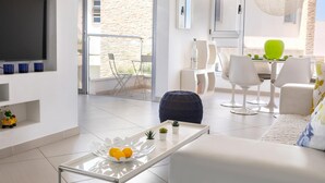 A modern living room with a flat-screen TV, a glass coffee table with oranges and small plants, white sofas, and a dining area with white chairs and a green chair. - Protaras Pallini Seaside Suite (Paralimni)