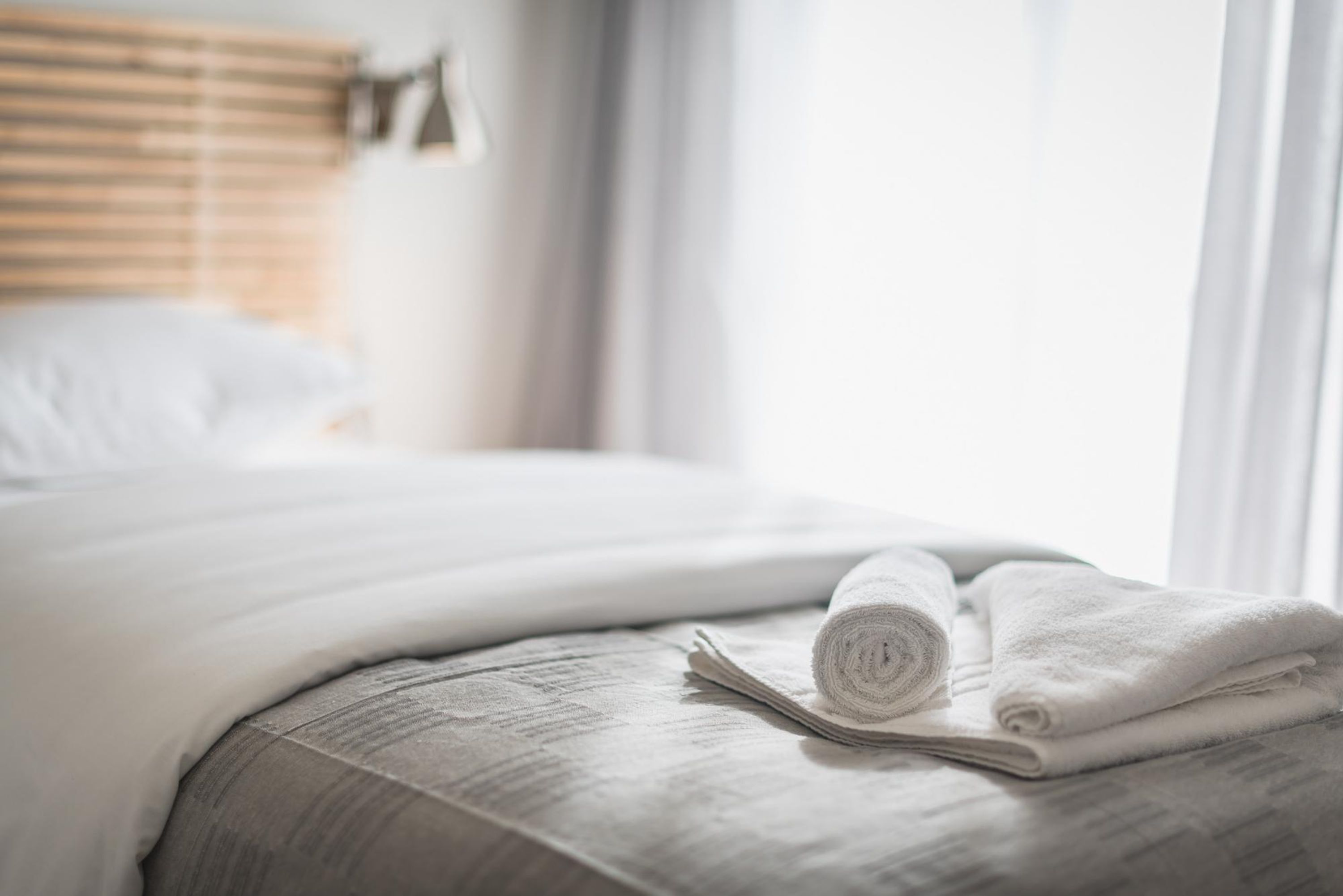 A neatly made bed with a rolled-up towel and a book on the side, in a room with a window and blinds.