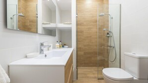 A modern bathroom with a white sink, mirror, and toilet. It features a glass shower enclosure and wooden flooring. - Fresh and Clean apt for 6 in Koukaki (Athens)