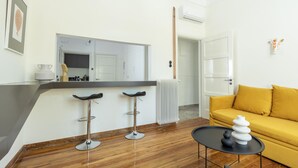 A modern living room with a yellow sofa, a black coffee table with decorative vases, a kitchen area with a large mirror, and a white radiator. - An Outstanding 3 Bdrm Apartment in the Heart of Athens (Athens)