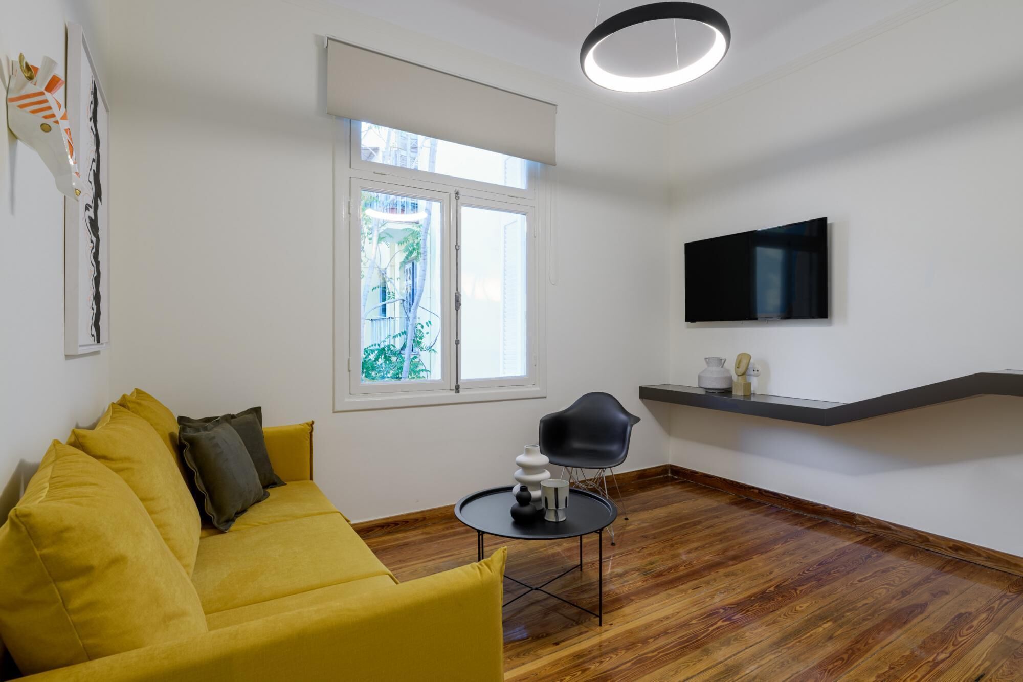 Una sala de estar moderna con un sofá amarillo, una silla negra, una mesa redonda, un televisor en la pared y un ventanal con vistas a la vegetación.