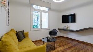 A modern living room with a yellow sofa, a black chair, a round table, a TV on a wall, and a window with a view of greenery. - An Outstanding 3 Bdrm Apartment in the Heart of Athens (Athens)