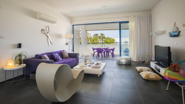 A modern living room with a purple sofa, a unique white chair, a coffee table, and a TV. - Aster Suite in Protaras (Paralimni)