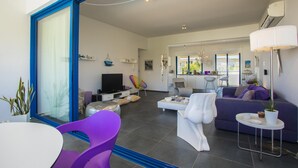 A modern living room with a large TV, a sofa, a dining table, and a chair. - Aster Suite in Protaras (Paralimni)