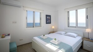 A bedroom with a bed, two bedside tables, a desk, and a window with a view of the sea. - Mackenzie Zoe Seafront Suite (Larnaca)