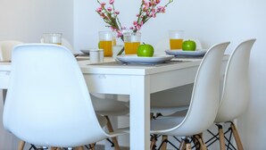 A dining table with white chairs, a vase with pink flowers, two green apples, and glasses of orange juice. - Mackenzie Zoe Seafront Suite (Larnaca)