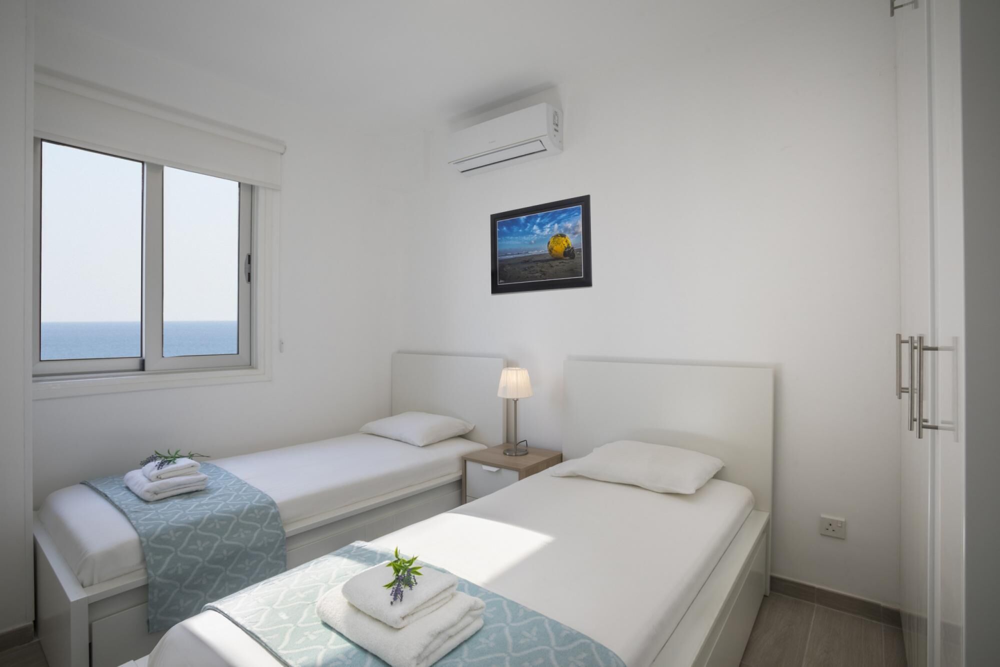 A hotel room with two beds, a window with a view of the sea, an air conditioner, and a framed picture on the wall.
