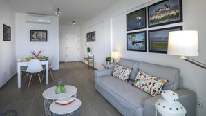 A modern living room with a grey sofa, a dining table with chairs, and a small round table with a green candle. - Mackenzie Zoe Seafront Suite (Larnaca)