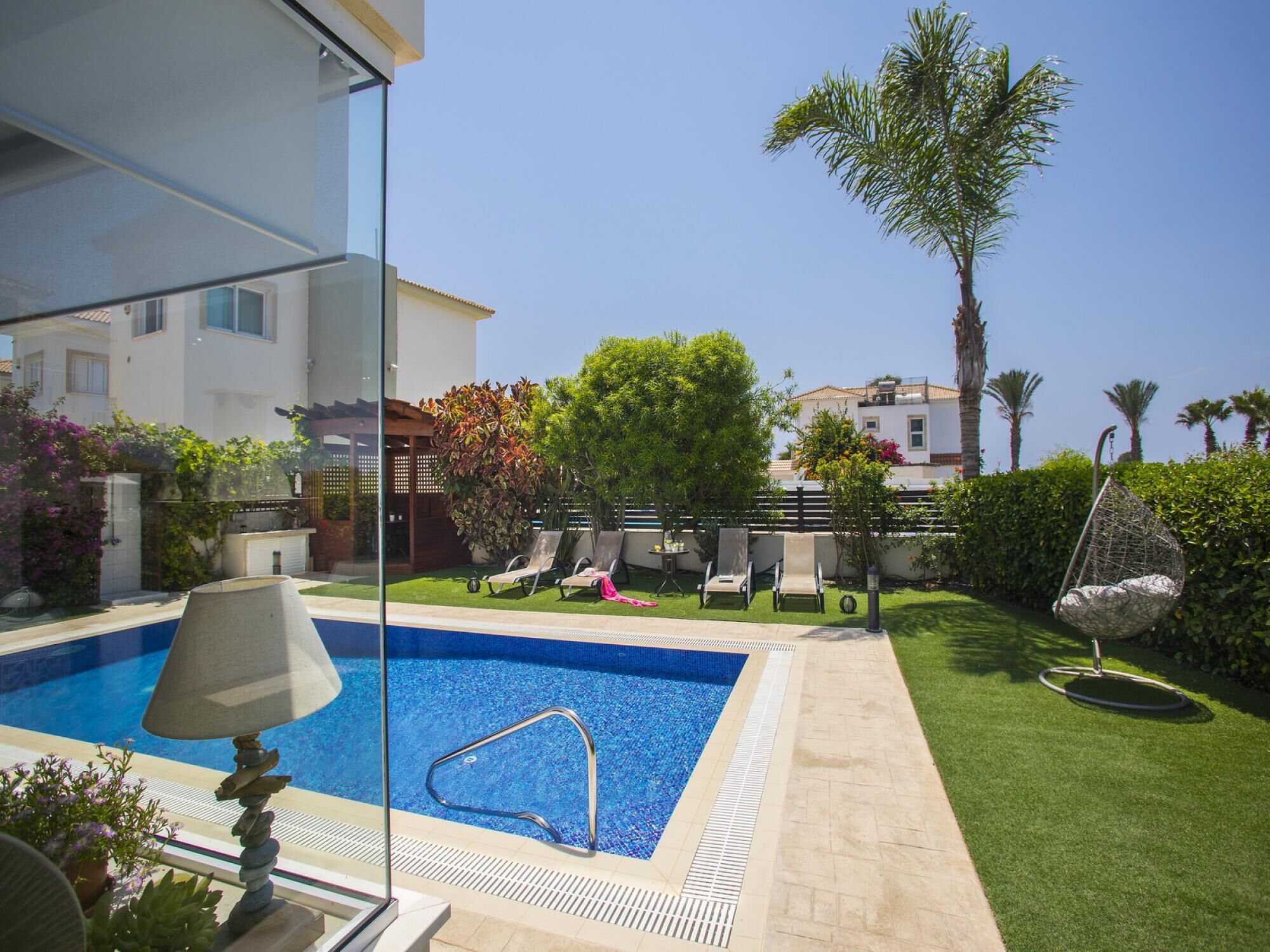 A swimming pool with a clear blue water, surrounded by a well-maintained lawn, a hanging chair, and a view of buildings in the background.