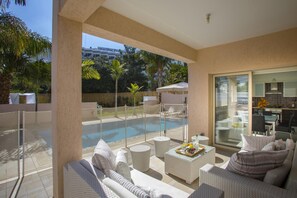 A modern outdoor living area with a pool, comfortable seating, and a glass door leading to a kitchen. - Protaras Villa Michelle 10 (Paralimni)