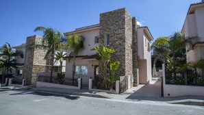 Une maison moderne à deux étages, dotée d’une façade en pierre, d’une entrée sécurisée par un portail et d’un jardin bien entretenu avec des palmiers.