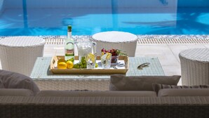 Un coin aménagé au bord de la piscine, avec un plateau contenant des citrons, des raisins et une bouteille de Sanjivi, ainsi qu’un verre d’eau et une carafe. L’ensemble est complété par des meubles en osier et une vue sur la piscine.