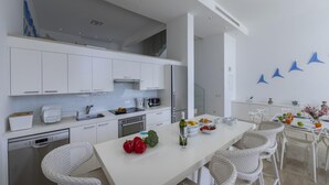 A modern kitchen with white cabinetry, stainless steel appliances, and a dining area with wicker chairs and a white table. - Protaras Villa Kalena (Paralimni)
