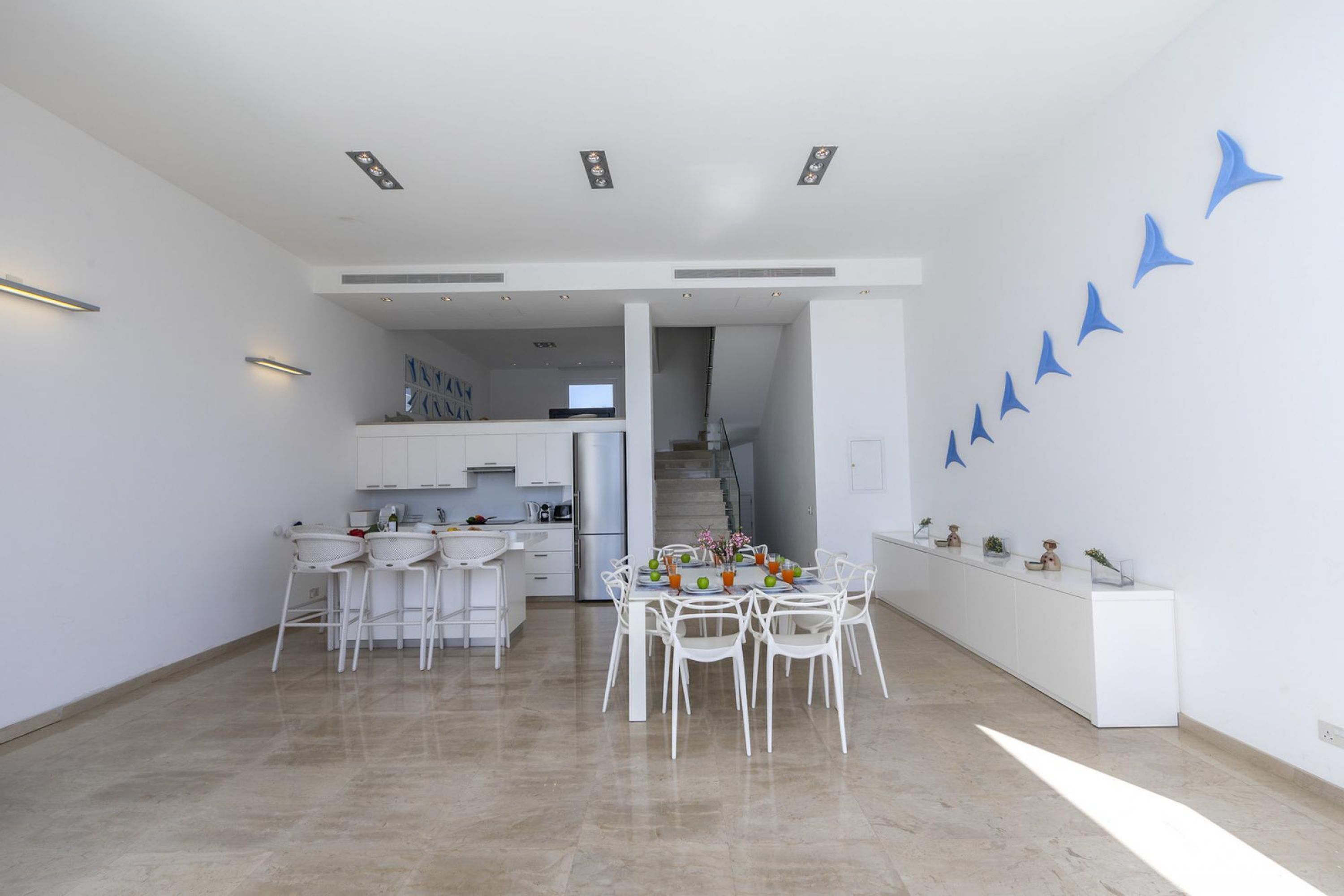 Una cocina moderna con armarios blancos, un comedor con sillas blancas y una escalera que sube a otro nivel.